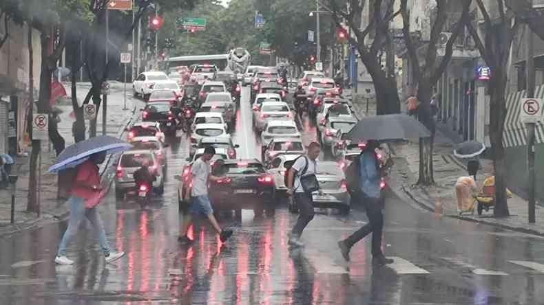 Imagem mostra quatro pessoas atravessando a rua na faixa de pedestres. Eles usam guarda-chuvas