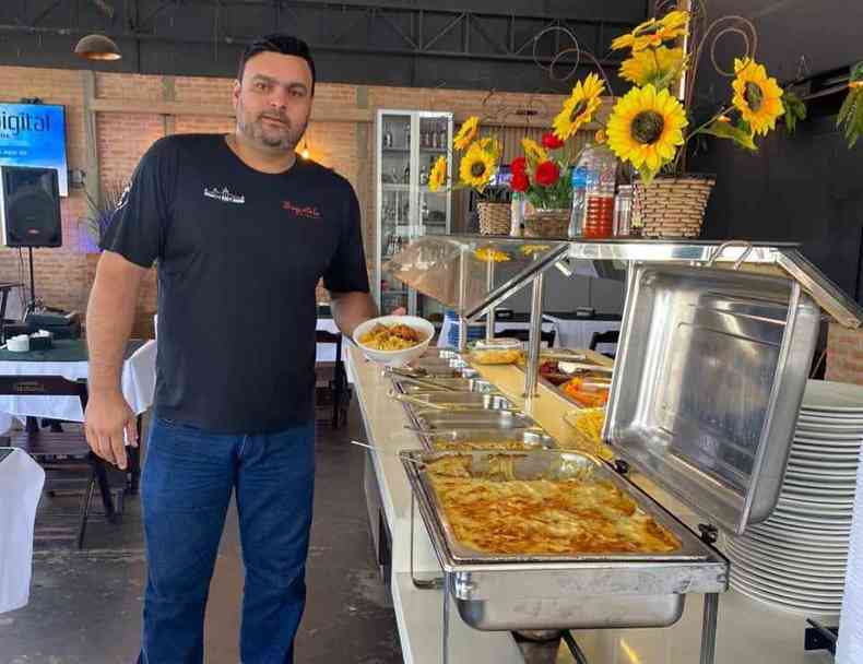 Homem est em p, vestindo cala jeans e blusa preta. Ele segura uma marmita de almoo com a mo esquerda