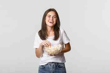 menina alegre comendo pipoca, est de blusa branca e cala jeans,  branca e possui cabelos castanhos