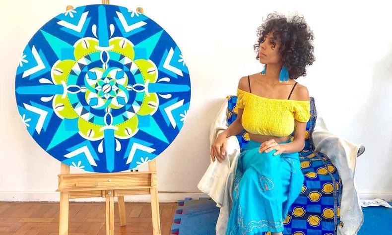 Pintora negra Bella Na est sentada e olha para mandala nas cores azul, branco e verde