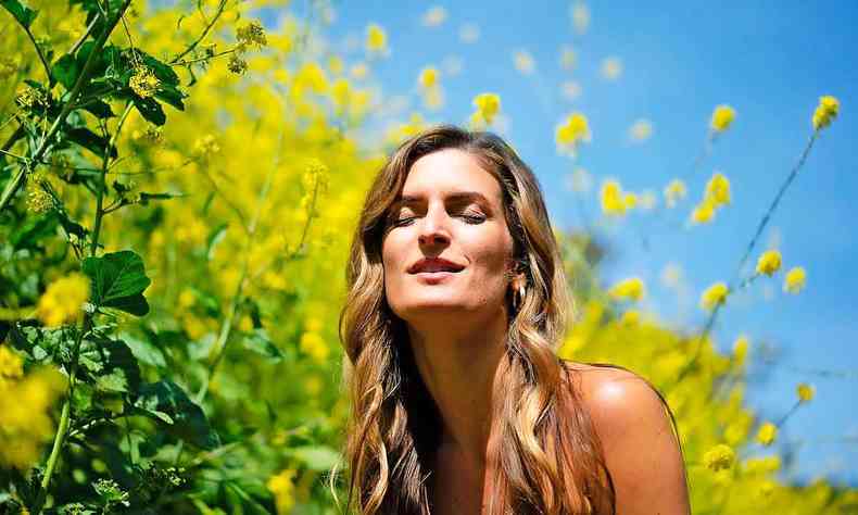 A cantora Daniela Soledade, de olhos fechados, ao ar livre, cercada de vegetação e exposta ao sol