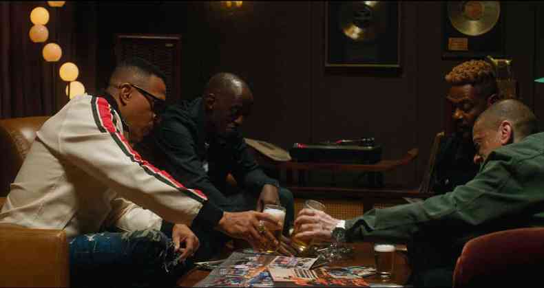 Os rappers Mano Brown, Ice Blue, Edi Rock e KL Jay esto sentados em volta de mesa, segurando copos de cerveja e segurando fotografias da banda