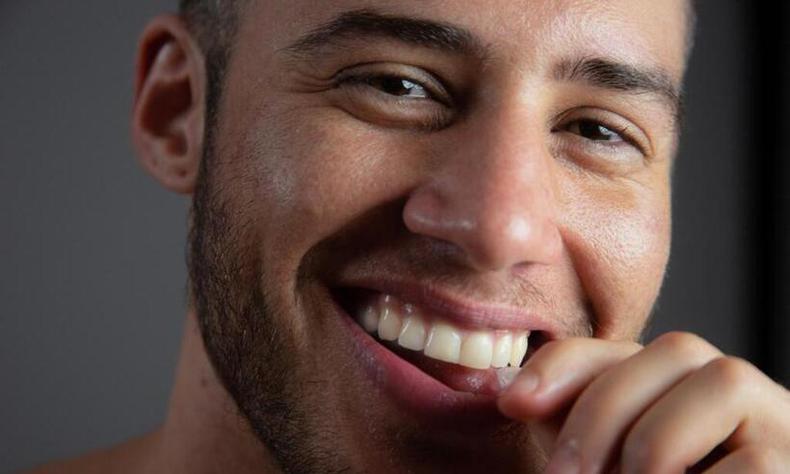 Foto do rosto de Paulo Vaz, um homem trans branco de cabelos e barba castanhos. Na imagem, ele sorri.