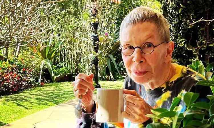 Rita Lee est com os cabelos grisalhos raspados e segura uma caneca e uma colher. Ao fundo, folhagens em um dia ensolarado