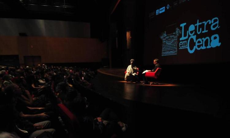 Conceio Evaristo, uma mulher preta de cabelos grisalhos presos no topo da cabea, e o jornalista Jos Eduardo Gonalves, um homem branco de cabelos grisalhos, esto iluminados no palco do Teatro. Na frente deles, uma grande plateia.