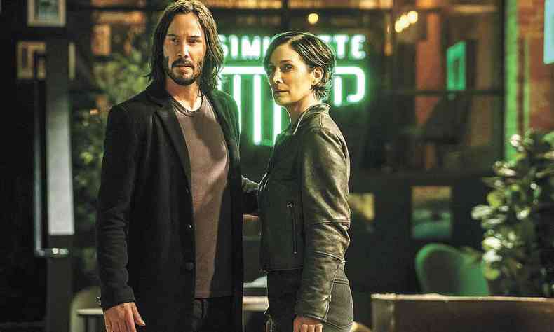 Keanu Reeves e Carrie-Anne Moss, preocupados, olham para a frente em cenrio escuro, com neon verde ao fundo, no filme 'Matrix resurrections'