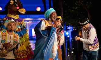 Lia de Itamarac usa turbante azul, brincos, vestido colorido e manto azul em cena de baile do menino deus