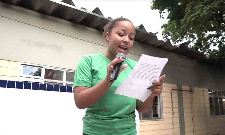 Menina negra usando uma blusa verde, recitando poema em microfone e segurando uma folha de papel