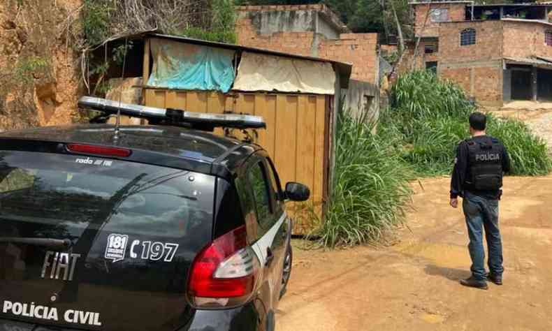 parte traseira de viatura da Polcia Civil aparece ao lado esquerdo. Policial em p  direita e ao fundo da imagem. No centro da imagem aparecem casas. 