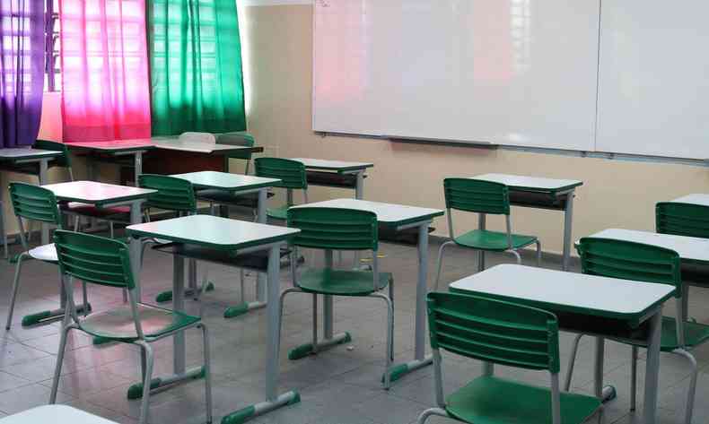 Sala de aula vazia, vista do fundo da sala. Cadeiras na cor verde e mesas brancas. Ao fundo, cortinas coloridas aparecem na janela