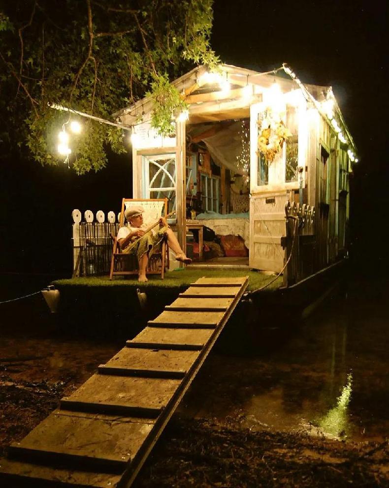 Foto mostra cabana em rio  noite iluminada por luzinhas e com uma mulher tocando banjo na varanda