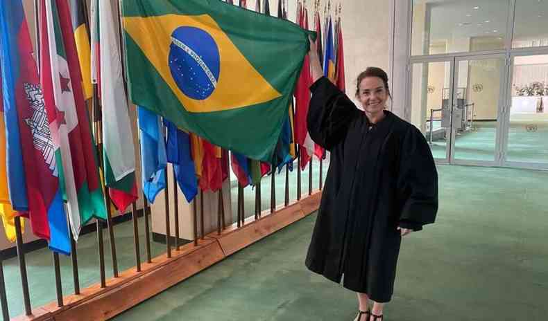 Martha, uma mulher branca de cabelos presos, vestindo toga, segura a bandeira do Brasil, pendurada perto de diversas outras bandeiras. 