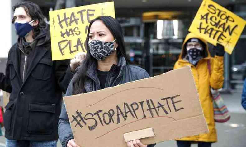 Uma mulher asitica amarela segura um cartaz com os dizeres 'STOP AAPI HATE'. Ela usa uma mscara preta e um casaco cinza. Atrs dela, h outros manifestantes