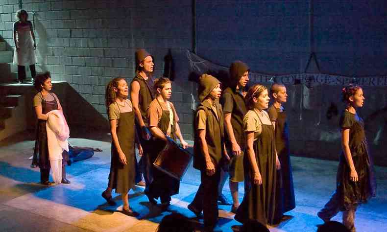 Vestidos com guardapó preto, atores e atrizes cantam e caminham pelo palco em cena que evoca procissão em 'Esta noite Mãe Coragem'