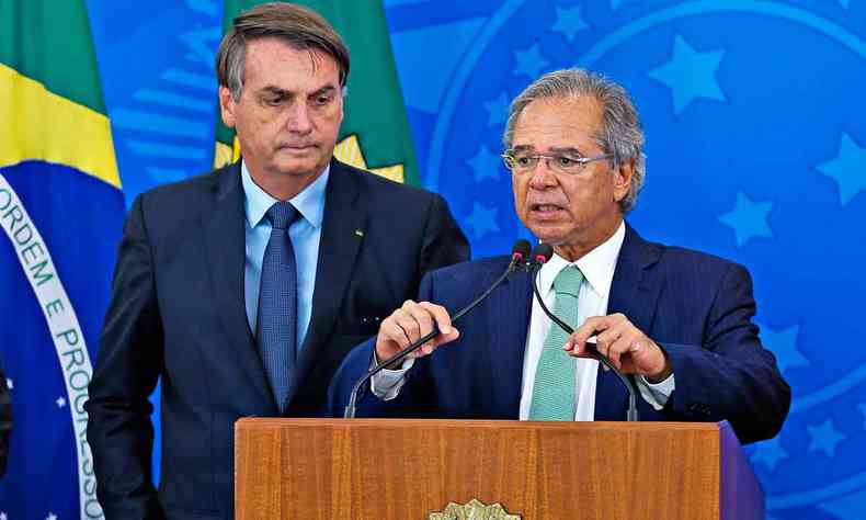 O ministro Paulo Guedes fala ao microfone, observado pelo presidente Jair Bolsonaro 