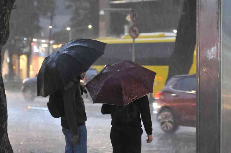 Na foto  possvel ver duas pessoas de costas segurando guarda-chuvas, elas esto atravessando a rua. 