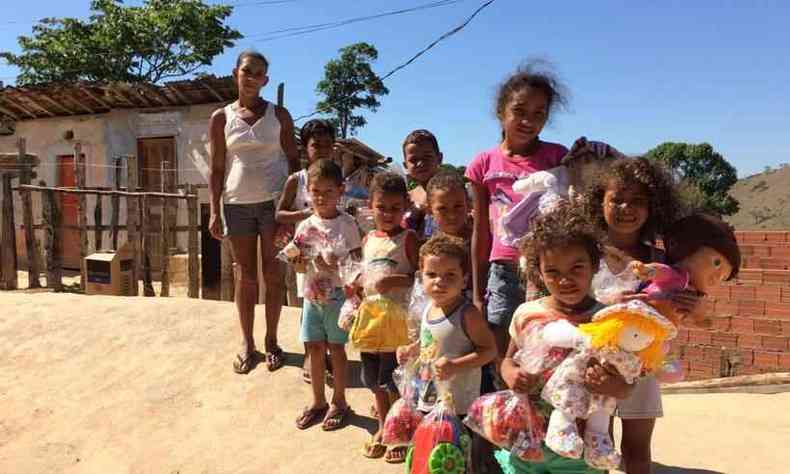 Uma mulher com várias crianças segurando brinquedos e alimentos doados, em frente a uma casa simples