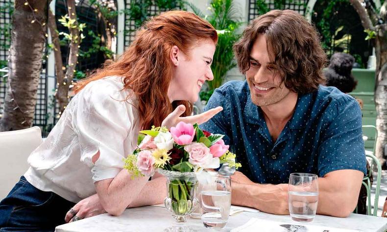 Sentados  mesa decorada com jarro de flores, os atores Rose Leslie (Clare) e Theo James (Henry) sorriem
