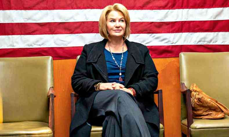 A atriz Renée Zellweger sentada numa cadeira com a bandeira dos EUA ao fundo olha para o lado em cena de 'The thing about Pam'