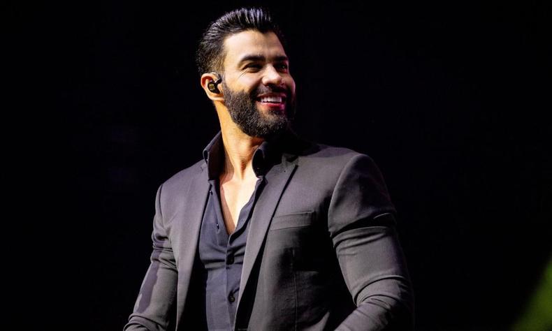 Astro do sertanejo, Gusttavo Lima se apresentará em Ipatinga, no Vale do Aço, em Minas Gerais, em 18 de novembro(foto: Gusttavo Lima/Site oficial/Divulgação) Gusttavo Lima sorrindo, todo de preto. Canhão de luz sobre ele. Fundo preto na imagem
