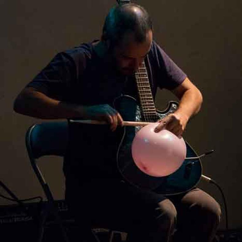 sentado de cala marrom e camisa preta, com a cabea abaixada sobre a guitarra, Matthias Koole pressiona um balo contra o instrumento