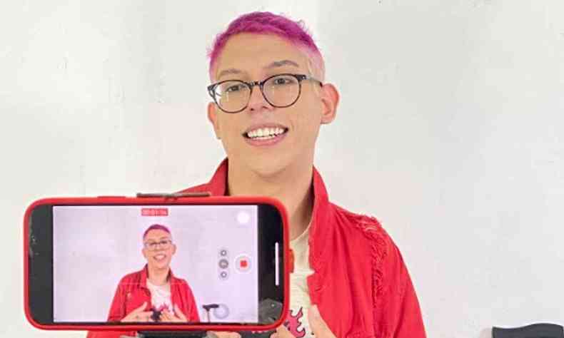 Ivan, com os cabelos rosa pink, usando uma blusa branca e uma jaqueta vermelha, est em frente ao celular, onde podemos ver sua imagem sendo capturada