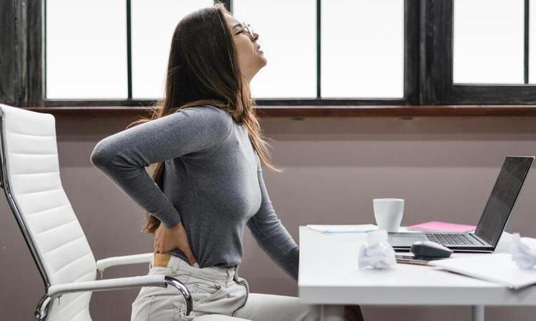 mulher no escritrio, sentada, leva a mo nas costas e tem expresso de dor