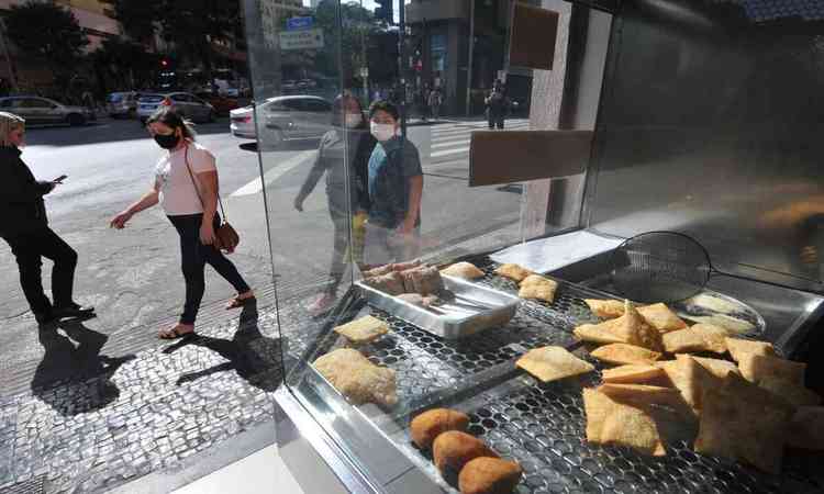 pessoas passando em frente a uma lanchonete, salgados podem ser vistos 