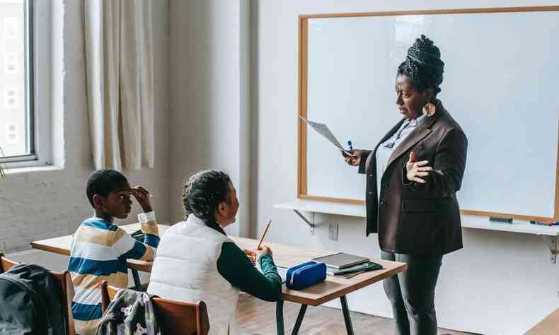 Sala de aula com uma professora negra e dois alunos sentados  sua frente