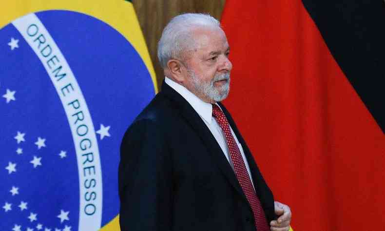 Lula. Ele  um homem branco de cabelos branco. Ao fundo a bandeira do Brasil 