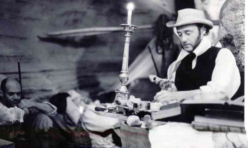 Cena do filme O homem de Lagoa Santa traz homem ao fundo, sentado no cho, e outro homem em primeiro plano,  frente de uma vela, sentado e mexendo em objetos sobre a mesa