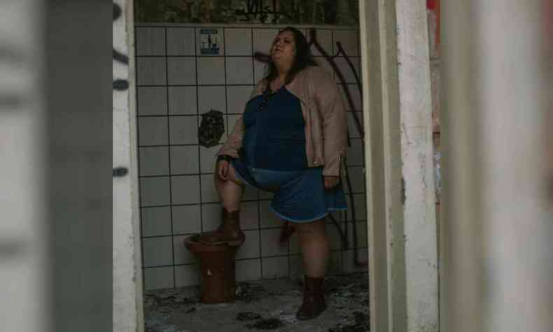 Jéssica, uma mulher gorda, se encontra em um banheiro sujo e degradado, apoia um dos pés na privada em posição de orgulho com o queixo erguido