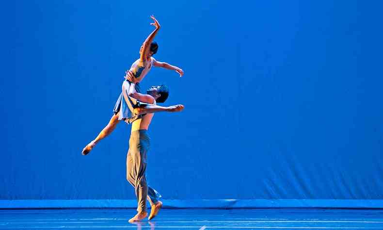 Bailarino ergue bailarina no palco, sob fundo azul. Ambos esto vestidos de branco
