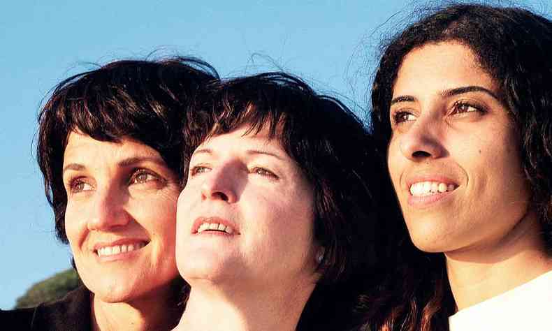 As cantoras Eveline Hecker, Georgiana de Moraes e Camilla Dias fotografadas lado a lado, sob o céu azul 