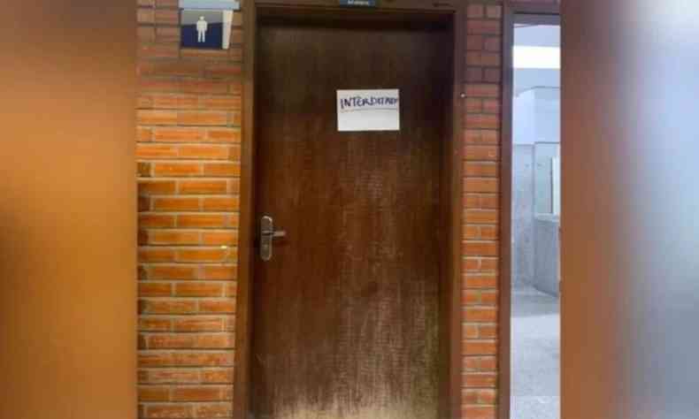 Imagem mostra porta de madeira de banheiro masculino da Faculdade de Comunicao da Universidade de Braslia. Ao lado, parede  feita de pequenos tijolos retangulares. 