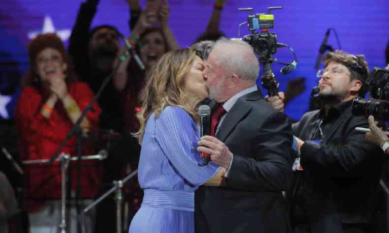 Janja, de vestido azul claro, beijando Lula em palco de evento