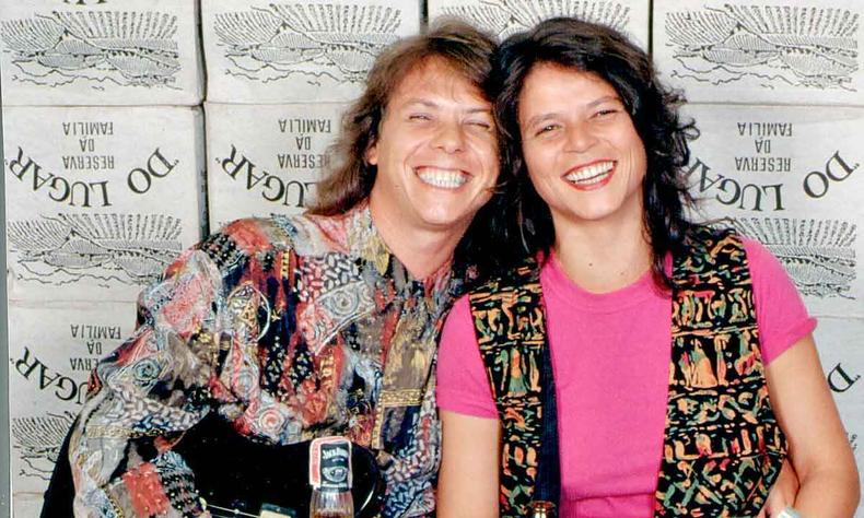 Victor Biglione e Cassia Eller sorriem diante de mesa de boteco, com copos de bebida e garrafa de usque