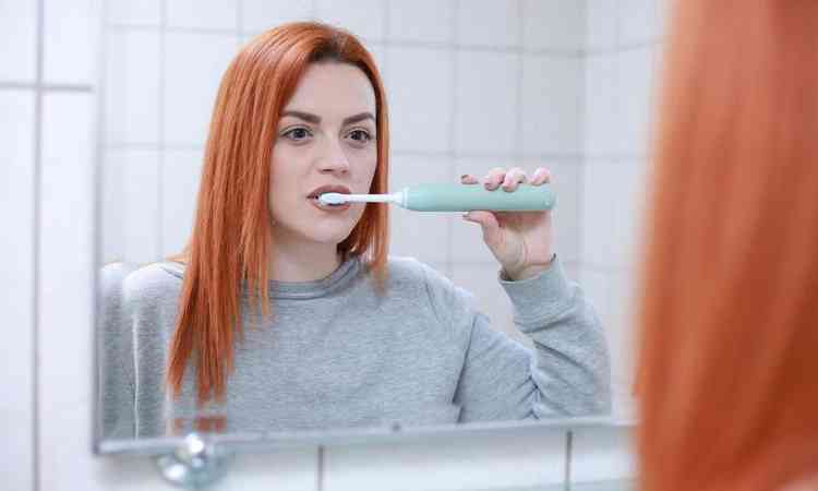 mulher de cabelo ruivo escova os dentes de frente para um espelho usando uma escova eltrica