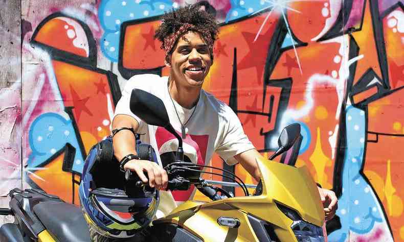 O ator Cau Campos sentado em uma moto, com parede grafitada ao fundo 