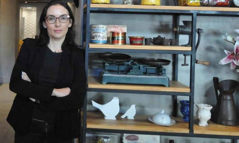 Carolina Moretzsohn olha para a câmera encostada em estante com objetos antigos, como balança, latas e objetos de cozinha
