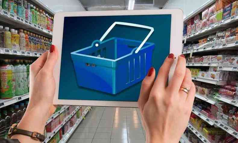 mulher segura tablet com imagem de uma cesta de supermercado diante de um corredor cheio de produtos 