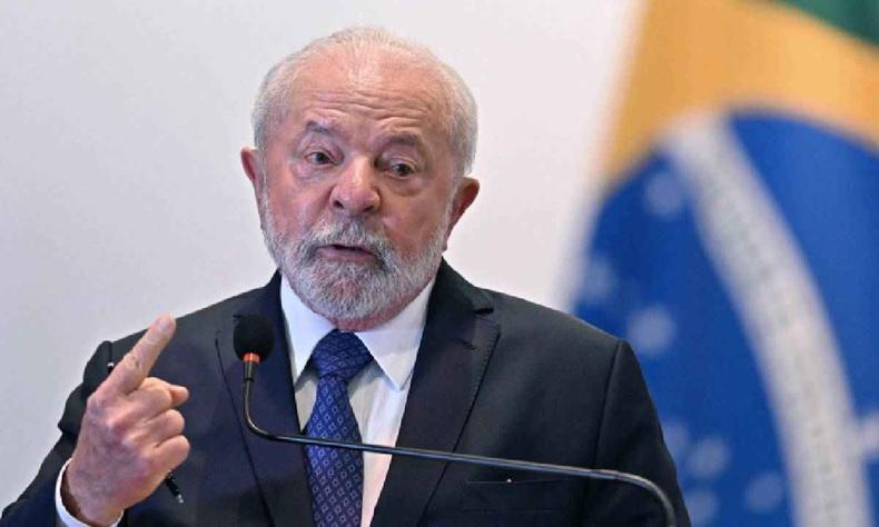 Lula, de terno preto, camisa branca e gravata azul falando ao microfone com uma bandeira do Brasil ao fundo, desfocada