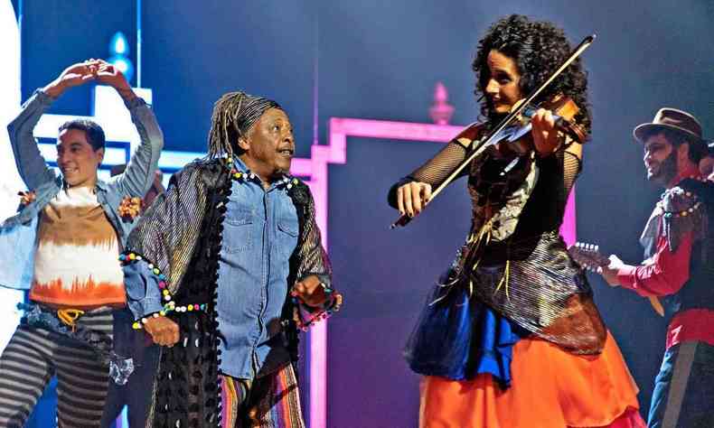 Tizumba canta no palco e olha para mulher tocando rabeca