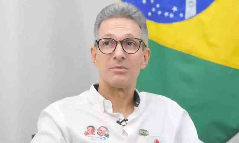 Romeu Zema sentado, de camisa branca, com a bandeira do Brasil ao fundo e um adesivo dele com o ex-presidente Jair Bolsonaro