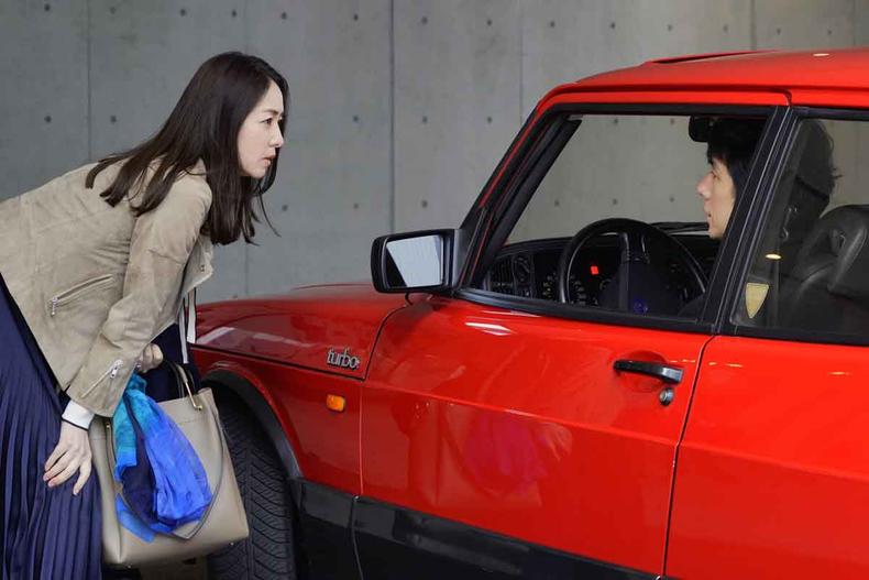 Atriz Reika Kirishima, com a mo no joelho, se agacha para falar com ator Hidetoshi Nishijima, sentado ao volante, dentro de carro vermelho, em cena de drive my car