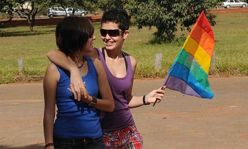 Duas mulheres se olham, sorriem e levam uma bandeira com as cores do arco-ris
