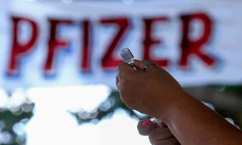 aplicação de vacina sendo preparada com faixa ao fundo, com a palavra Pfizer em vermelho