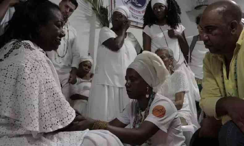 Imagem mostra integrante de comunidade quilombola em um ritual religioso. Sete pessoas, vestidas de branco, aparecem em volta de Me Efignia, fundadora da comunidade, que est sentada