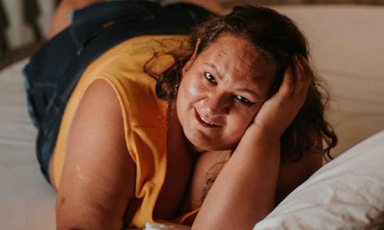 J�ssica, uma mulher gorda, deitava em uma cama com o rosto apoiado na m�o esquerda, usa blusa amarela e cal�a preta. ela sorri para a foto