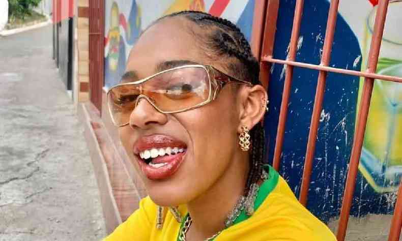 Nzambi  uma mulher preta e usa tranas em seus cabelos e um culos de sol com lentes grandes e marrons. Usa um brinco dourado e uma camiseta da seleo brasileira de futebol
